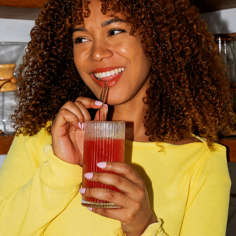 Lächelnde Frau in einem gelben Oberteil, die ein rotes Tavo LiquidCalm Liquid-Calm-Getränk trinkt und einen Stickpack in ihrer Küche hält.